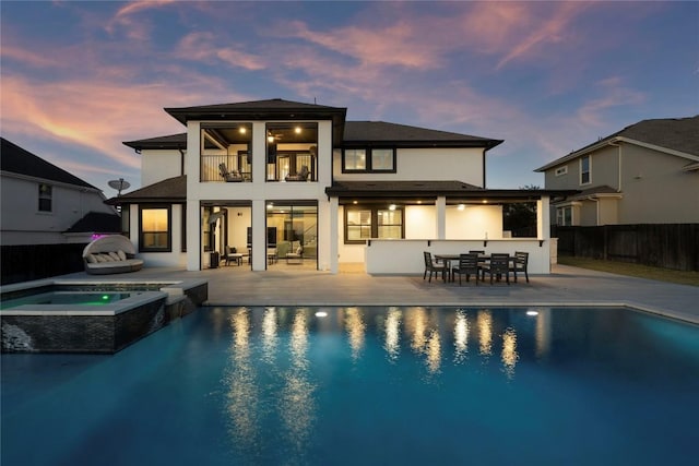 back house at dusk with a balcony, a swimming pool with hot tub, an outdoor bar, and a patio area