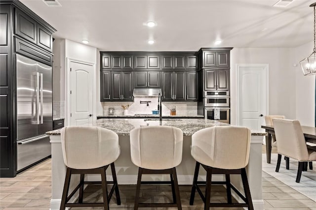 kitchen featuring appliances with stainless steel finishes, hanging light fixtures, tasteful backsplash, light stone countertops, and a center island with sink