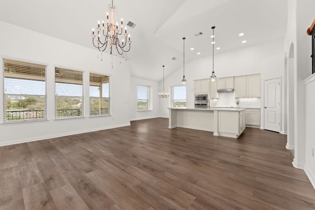 kitchen featuring pendant lighting, stainless steel microwave, high vaulted ceiling, an island with sink, and a notable chandelier