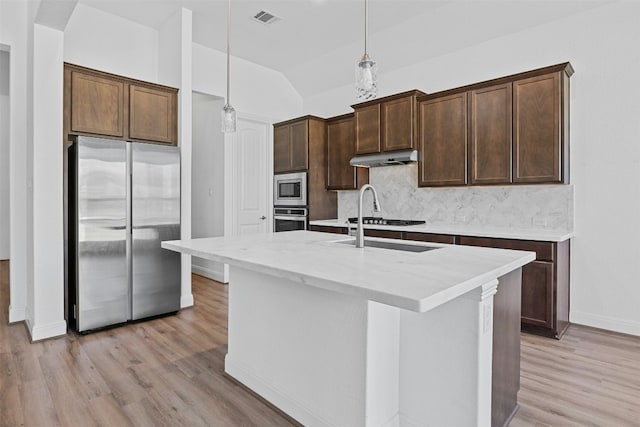 kitchen featuring appliances with stainless steel finishes, decorative light fixtures, an island with sink, dark brown cabinets, and light hardwood / wood-style flooring