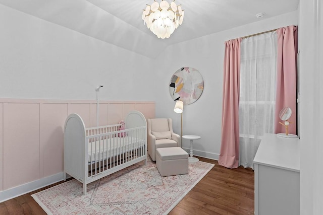 bedroom with a wainscoted wall, an inviting chandelier, vaulted ceiling, wood finished floors, and a nursery area