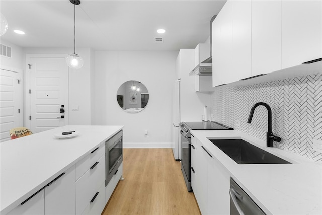 kitchen with appliances with stainless steel finishes, pendant lighting, white cabinetry, wall chimney range hood, and light wood-type flooring