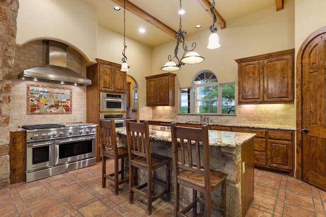 kitchen with wall chimney range hood, beam ceiling, stainless steel appliances, a center island, and light stone counters