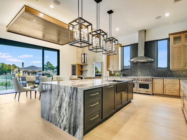 kitchen featuring pendant lighting, double oven range, wall chimney exhaust hood, light stone counters, and a large island with sink