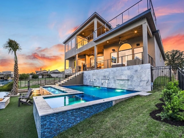 back house at dusk featuring ceiling fan, a balcony, a swimming pool with hot tub, and a yard