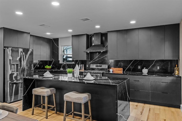 kitchen featuring a breakfast bar area, a center island, light wood-type flooring, appliances with stainless steel finishes, and wall chimney range hood