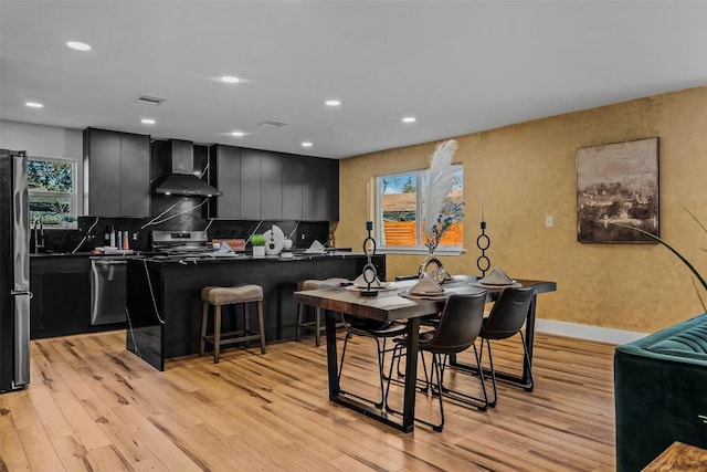 kitchen with wall chimney exhaust hood, tasteful backsplash, light hardwood / wood-style flooring, appliances with stainless steel finishes, and a kitchen breakfast bar