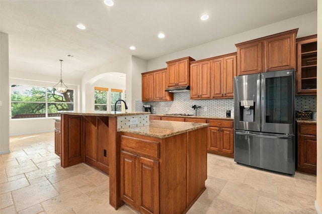 kitchen featuring stainless steel refrigerator with ice dispenser, light stone counters, hanging light fixtures, a kitchen island with sink, and backsplash