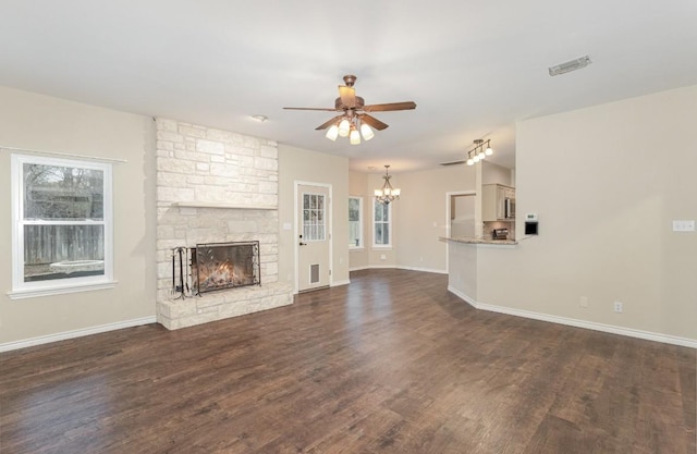 unfurnished living room with dark wood-type flooring, a stone fireplace, baseboards, and ceiling fan with notable chandelier