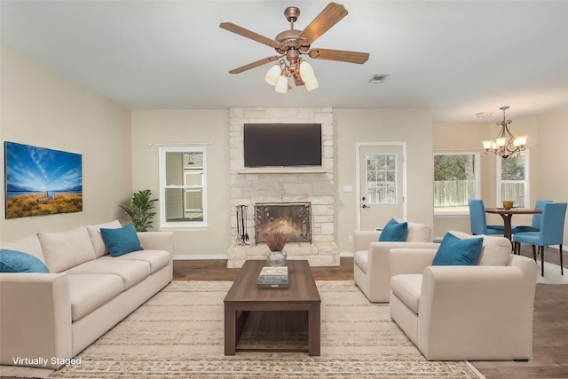 living room featuring ceiling fan with notable chandelier, visible vents, wood finished floors, and a stone fireplace