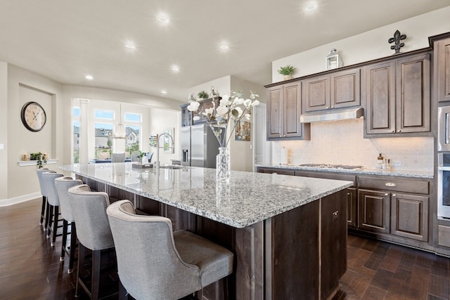 kitchen featuring light stone counters, dark wood finished floors, backsplash, a large island with sink, and under cabinet range hood