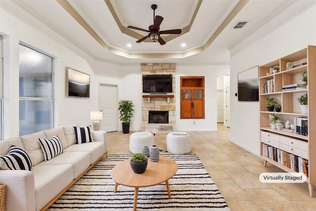 tiled living room featuring a tray ceiling, a stone fireplace, ornamental molding, and ceiling fan