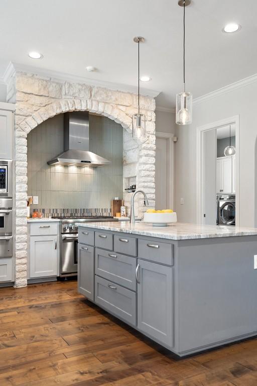 kitchen featuring gray cabinetry, light stone countertops, ornamental molding, washer / dryer, and wall chimney exhaust hood