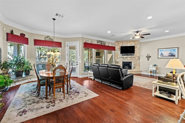 dining area with wood-type flooring, a stone fireplace, ceiling fan with notable chandelier, and crown molding