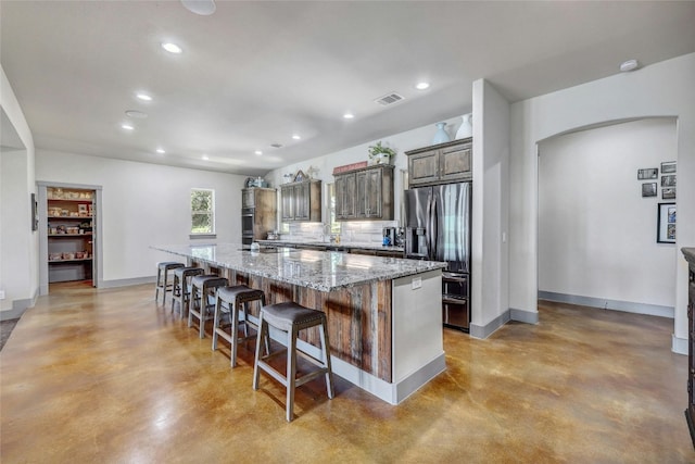 kitchen with a center island with sink, light stone countertops, a kitchen bar, stainless steel fridge with ice dispenser, and concrete floors