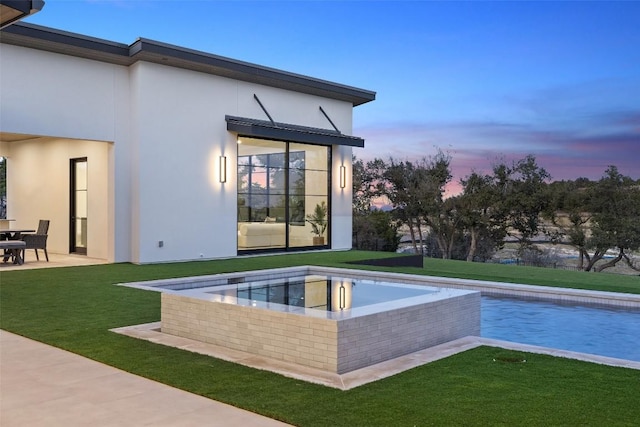 pool at dusk featuring a hot tub, a patio area, and a lawn