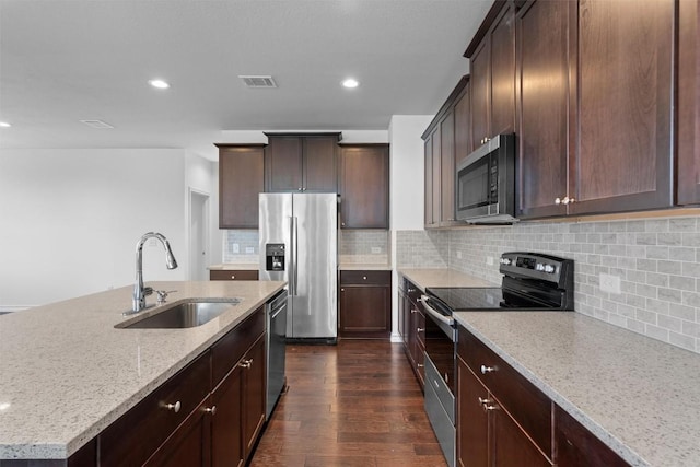 kitchen with sink, dark wood-type flooring, appliances with stainless steel finishes, a kitchen island with sink, and light stone countertops