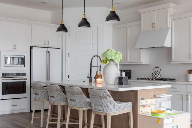 kitchen with premium range hood, appliances with stainless steel finishes, decorative light fixtures, white cabinets, and a center island with sink