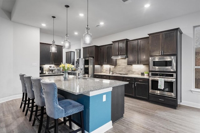 kitchen with light stone counters, tasteful backsplash, decorative light fixtures, appliances with stainless steel finishes, and an island with sink