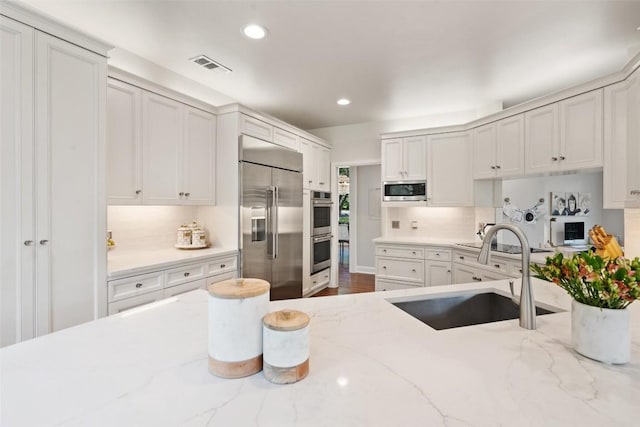 kitchen featuring sink, tasteful backsplash, built in appliances, light stone countertops, and white cabinets