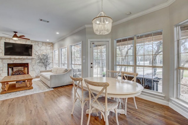 dining area with a stone fireplace, wood-type flooring, ornamental molding, and ceiling fan with notable chandelier