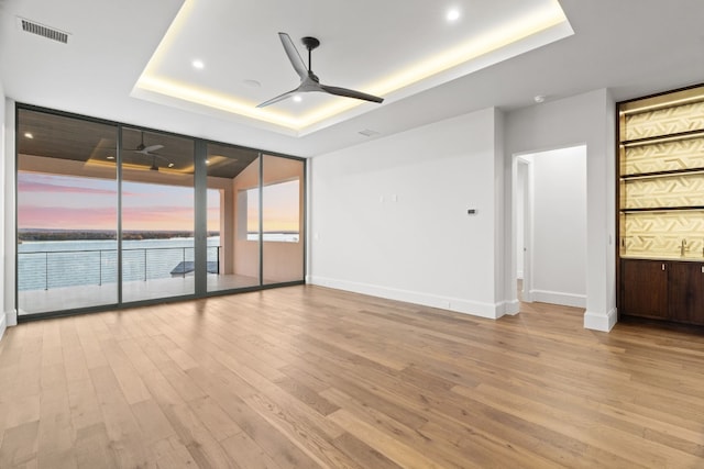 unfurnished room with a water view, light hardwood / wood-style flooring, a tray ceiling, expansive windows, and ceiling fan