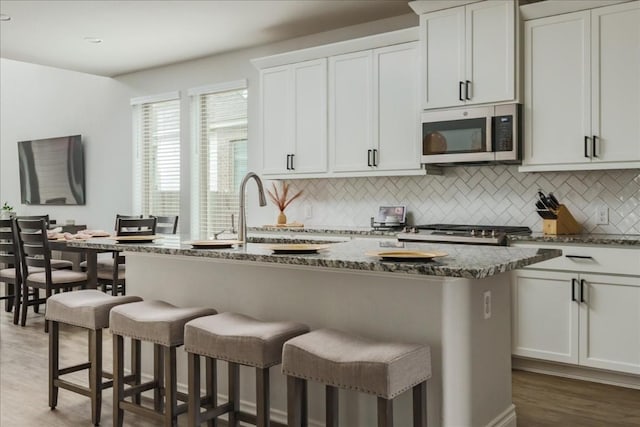 kitchen featuring an island with sink, dark stone countertops, white cabinets, decorative backsplash, and dark wood-type flooring