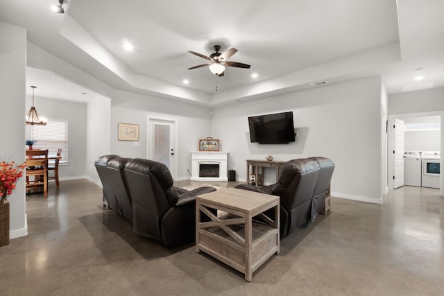 living room with independent washer and dryer, a tray ceiling, concrete flooring, and ceiling fan with notable chandelier