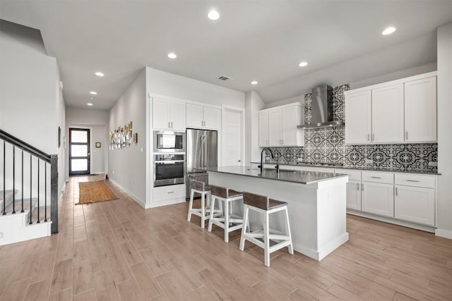 kitchen featuring an island with sink, white cabinets, a kitchen bar, stainless steel appliances, and wall chimney range hood