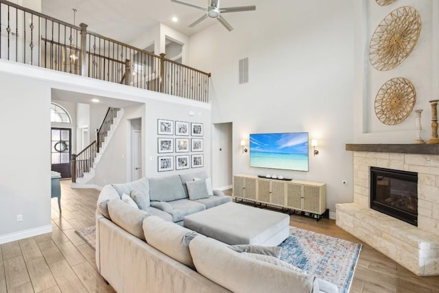 living room featuring ceiling fan, a stone fireplace, light hardwood / wood-style flooring, and a towering ceiling