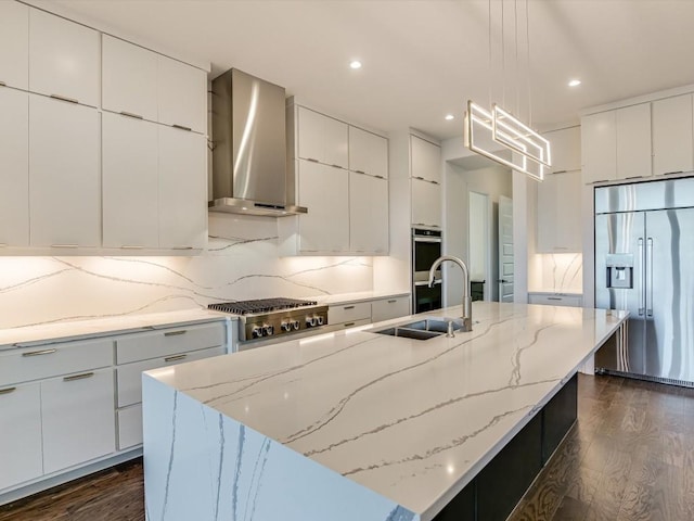 kitchen with appliances with stainless steel finishes, hanging light fixtures, white cabinets, a center island with sink, and wall chimney exhaust hood