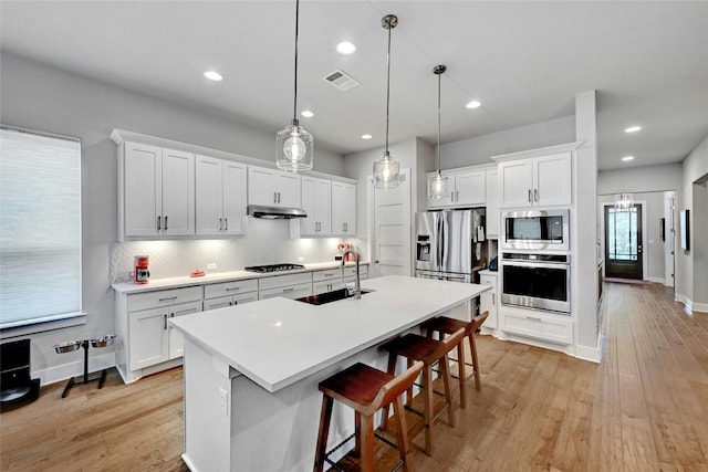 kitchen featuring appliances with stainless steel finishes, a center island with sink, white cabinets, and decorative light fixtures