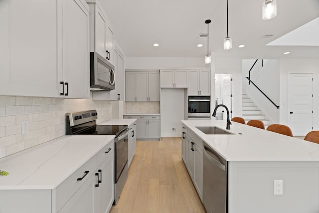 kitchen with sink, stainless steel appliances, light stone counters, an island with sink, and decorative light fixtures