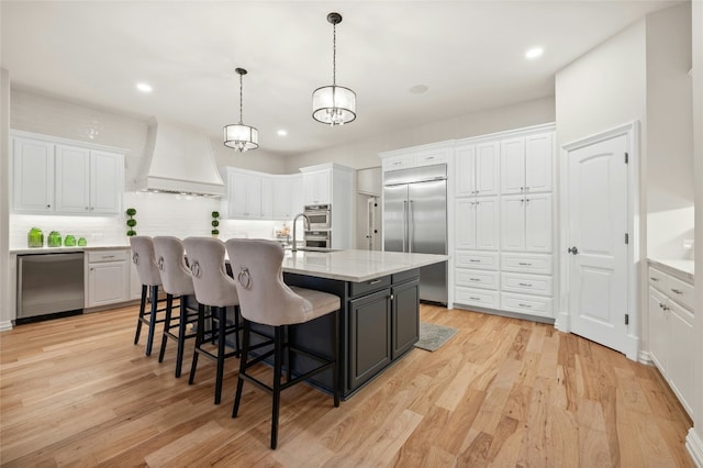 kitchen with white cabinetry, appliances with stainless steel finishes, custom range hood, pendant lighting, and a kitchen island with sink