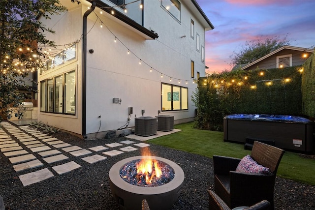 back house at dusk featuring a hot tub, a yard, cooling unit, and an outdoor fire pit