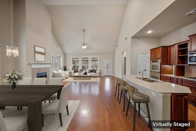 kitchen featuring ceiling fan with notable chandelier, high vaulted ceiling, a tiled fireplace, stainless steel appliances, and light hardwood / wood-style flooring