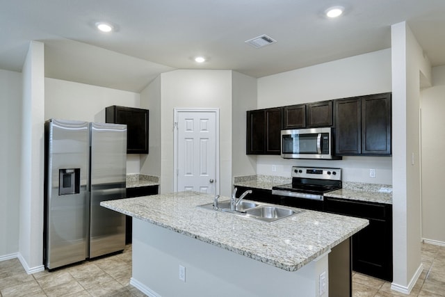 kitchen featuring appliances with stainless steel finishes, sink, a kitchen island with sink, dark brown cabinetry, and light stone countertops