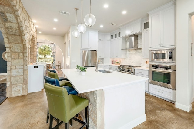 kitchen featuring an island with sink, appliances with stainless steel finishes, light countertops, wall chimney range hood, and a sink