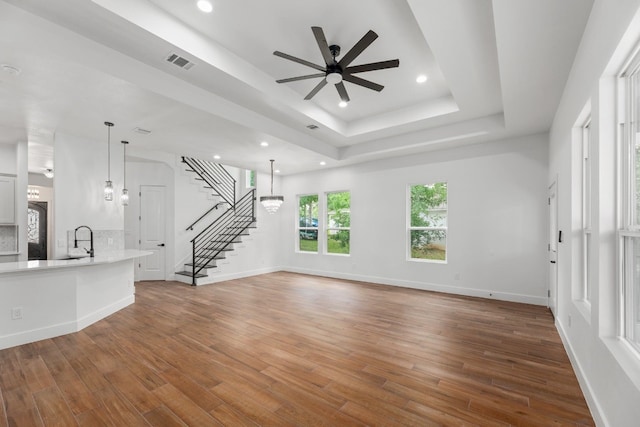 unfurnished living room with hardwood / wood-style floors, sink, ceiling fan with notable chandelier, and a raised ceiling