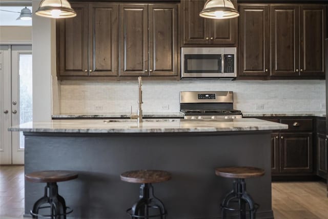 kitchen featuring a kitchen island with sink, stainless steel appliances, a kitchen breakfast bar, dark brown cabinetry, and light stone countertops