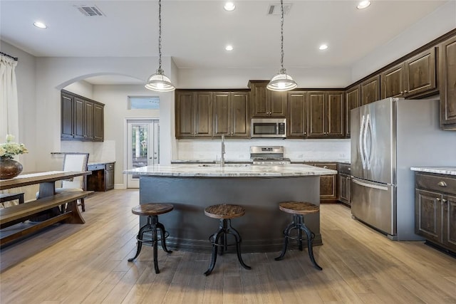 kitchen with appliances with stainless steel finishes, pendant lighting, a kitchen island with sink, light stone countertops, and dark brown cabinets