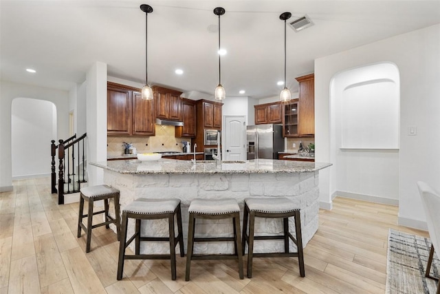 kitchen featuring light stone counters, hanging light fixtures, an island with sink, stainless steel appliances, and backsplash