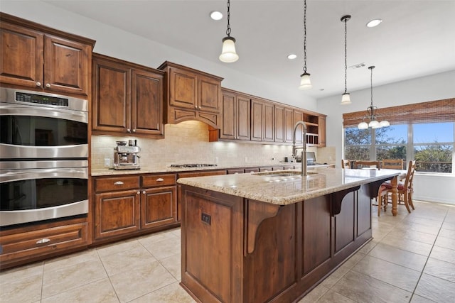 kitchen featuring sink, appliances with stainless steel finishes, a kitchen island with sink, hanging light fixtures, and light stone counters