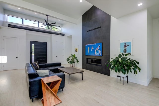 living room with ceiling fan, a tile fireplace, a high ceiling, and light wood-type flooring