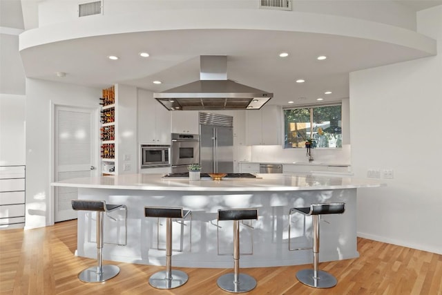 kitchen featuring white cabinetry, island exhaust hood, a kitchen bar, and built in appliances