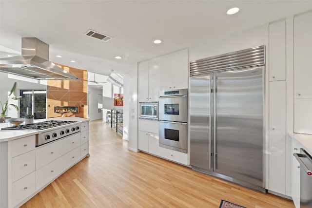 kitchen featuring built in appliances, light wood-type flooring, island exhaust hood, decorative backsplash, and white cabinets