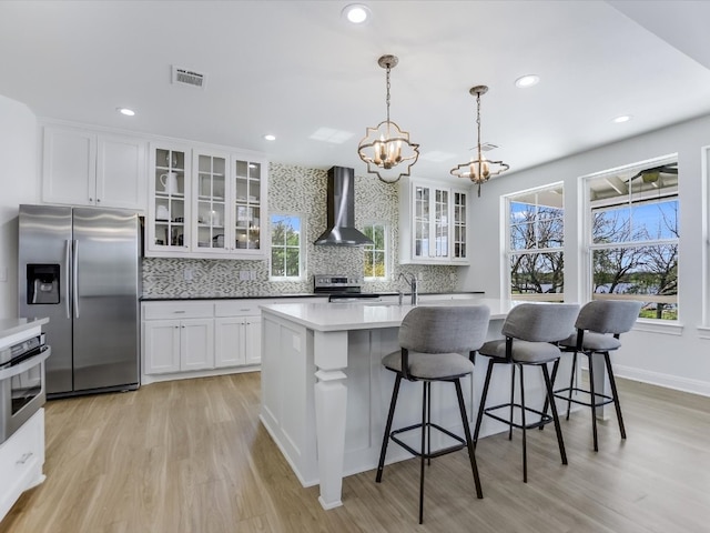 kitchen featuring wall chimney exhaust hood, a center island with sink, appliances with stainless steel finishes, pendant lighting, and white cabinets