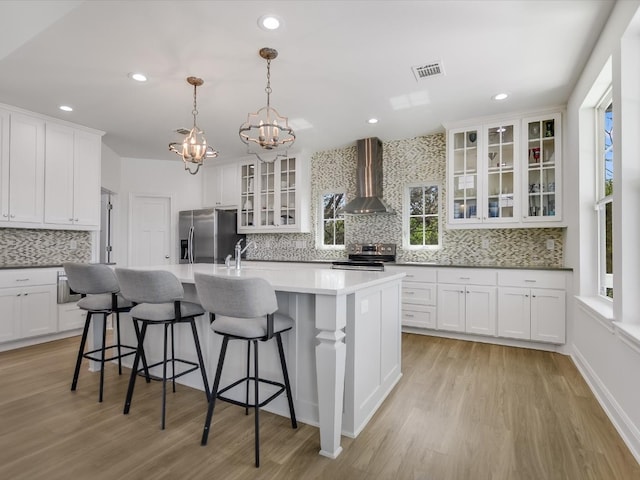 kitchen featuring wall chimney exhaust hood, white cabinetry, hanging light fixtures, a center island with sink, and appliances with stainless steel finishes