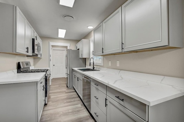 kitchen with appliances with stainless steel finishes, a skylight, sink, light hardwood / wood-style floors, and light stone counters