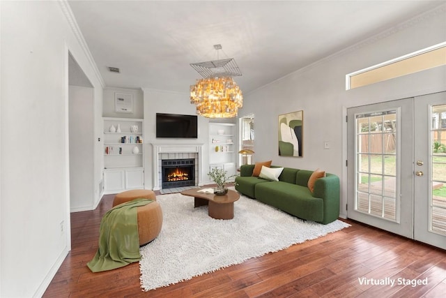 living room featuring a tile fireplace, built in features, wood-type flooring, ornamental molding, and a notable chandelier